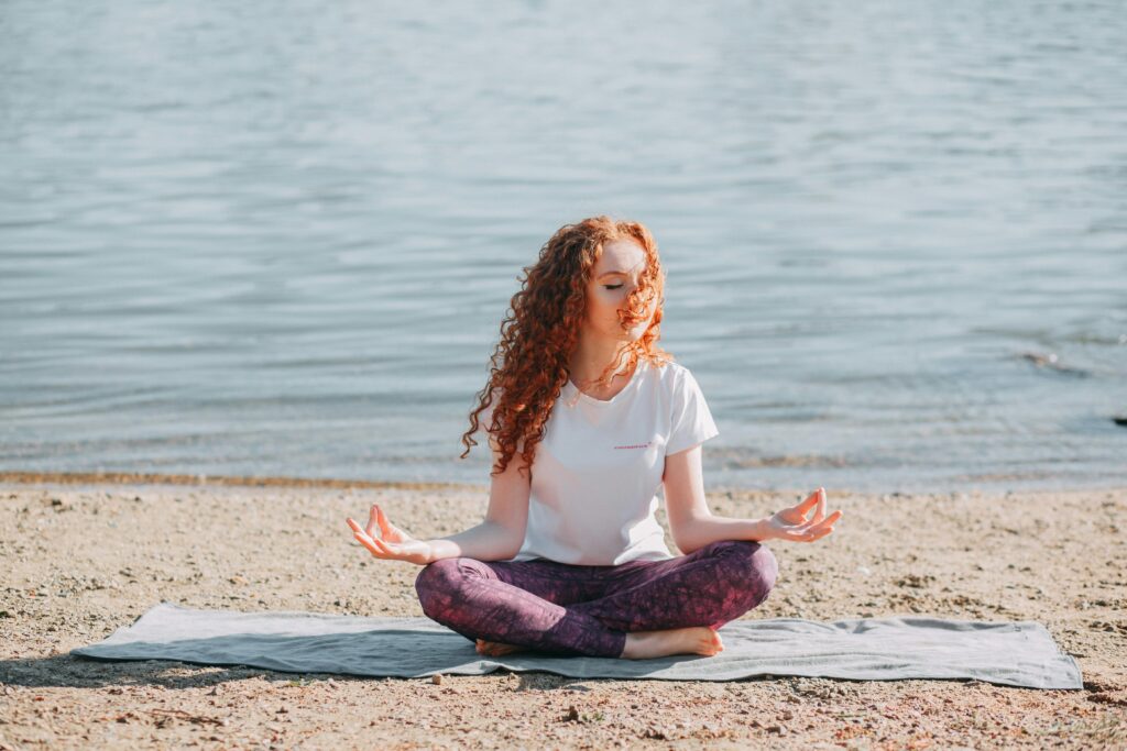 Journaling Made Me Better: 5 Powerful Ways It Transformed My Mental Health 3 A person with red curly hair is sitting in a meditative pose on a mat by the water, eyes closed and hands resting on their knees. This serene scene could represent the inner peace and improved well-being that might come from practices like journaling, suggesting that "journaling made me better".
